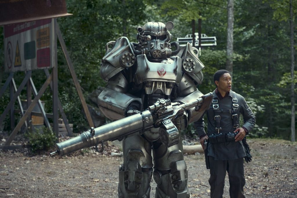 Still image from the Amazon post-apocalyptic drama series "Fallout" featuring Aaron Moten as Maximus standing behind Knight Titus in silver armor holding a machine gun. 
