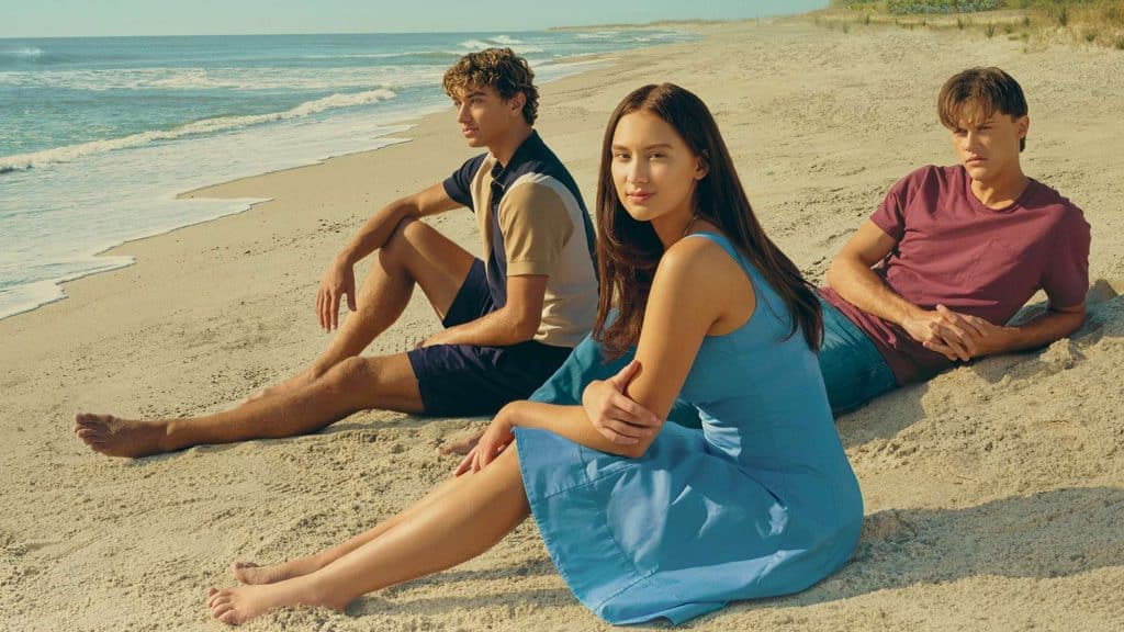 Promotional image from the Amazon romantic drama series "The Summer I Turned Pretty" featuring stars Lola Tung, Gavin Casalegno, and Christoper Briney sitting on a beach. 