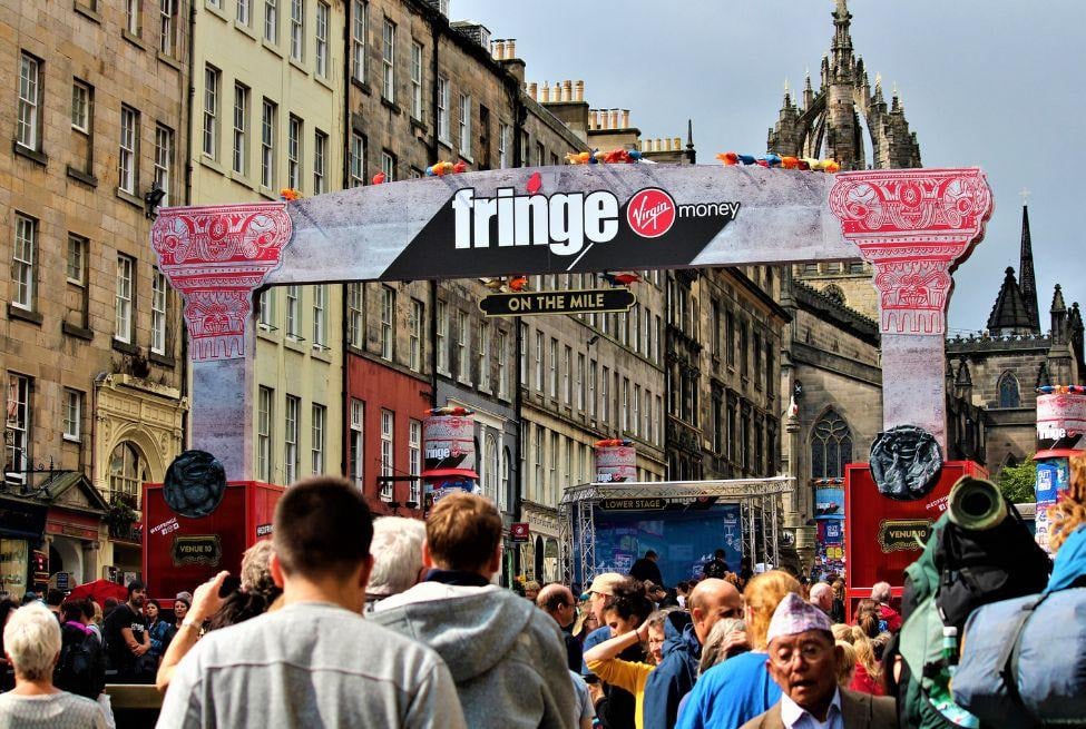 Photograph featuring a marquee for the Edinburgh Fringe Festival with pedestrians in the foreground and stone buildings in the background. 