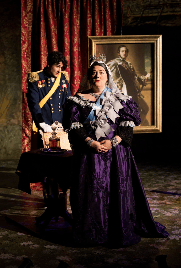 A still image featuring Prince Leopold in a military uniform and Queen Victoria in a crown and purple gown from the production of the musical "Alice, Formerly of Wonderland".