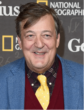 Photograph of actor and comedian Stephen Fry wearing a light blue suit jacket, maroon vest, dark red checked shirt, and a yellow tie with white dots. 