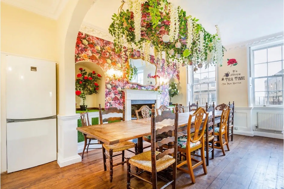 A dining room featuring a wood dining table, flower-themed wallpaper, and a plant installation hanging over the table from The Wonderland Townhouse in Bath, England. 