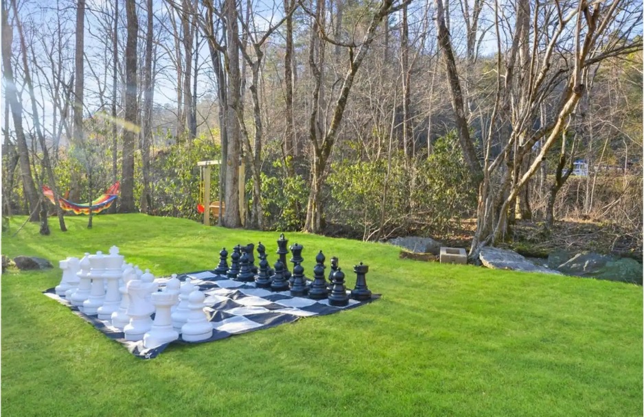 Backyard with a green lawn set against the woods featuring a large black and white chess set from the Alice's Queen Cottage Airbnb in Hayesville, North Carolina. 