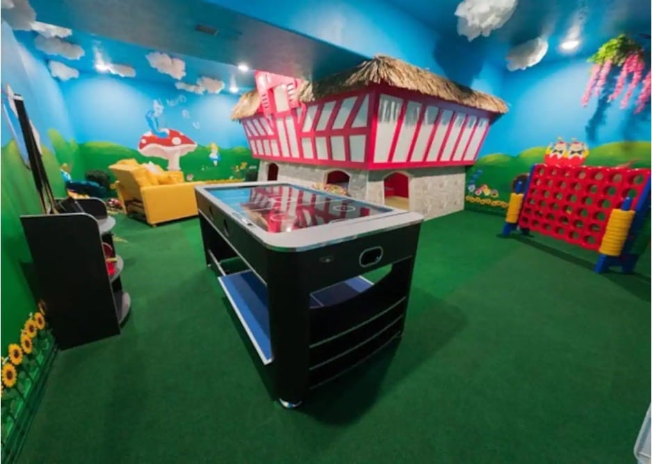 A playroom featuring an air hockey table, a large pink and white playhouse, and a blue and green landscape mural painted on the wall from the Alice in Wonderland Airbnb in Ozark, Missouri. 