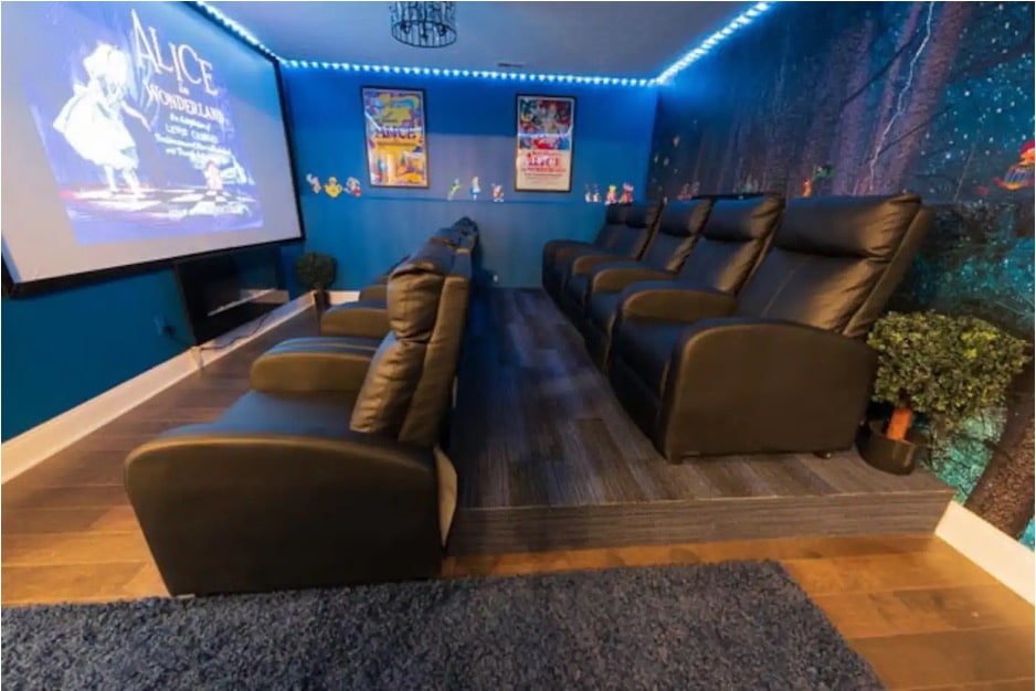 A screening room with black leather chairs, a nighttime forest-themed mural, and a projection screen from the Alice in Wonderland Airbnb in Ozark, Missouri. 