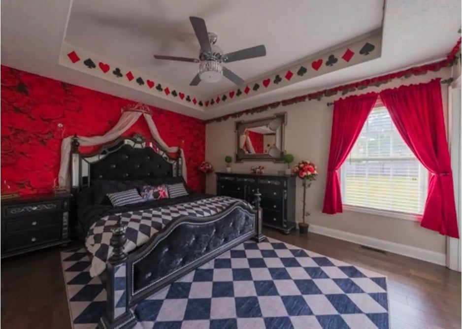 A bedroom with red wallpaper and curtains, card suit-themed decorations, and a blue and white diamond rug and comforter from the Alice in Wonderland Airbnb in Ozark, Missouri. 