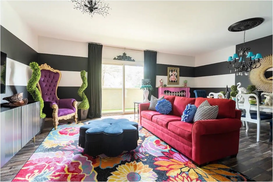 A living room with a red couch, flower-themed rug, purple armchair, and black-and-white striped wallpaper from the Alice's Palace Airbnb in St. Augustine, Florida. 