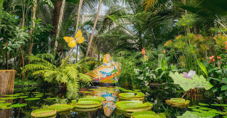 A small pool with lily pads surrounded by greenery featuring an "Alice in Wonderland" statue from the Wonderland: Curious Nature installation at the New York Botanical Garden. 