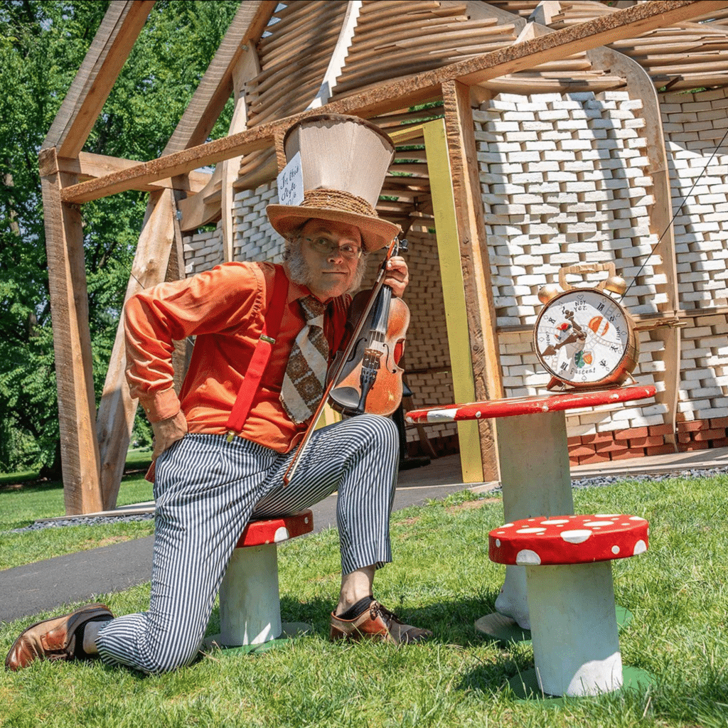 A photograph of an actor holding a violin and dressed as the Mad Hatter from Lewis Carroll's "Alice's Adventures in Wonderland" as part of the "Wonderland: Curious Nature" exhibition at the New York Botanical Garden.