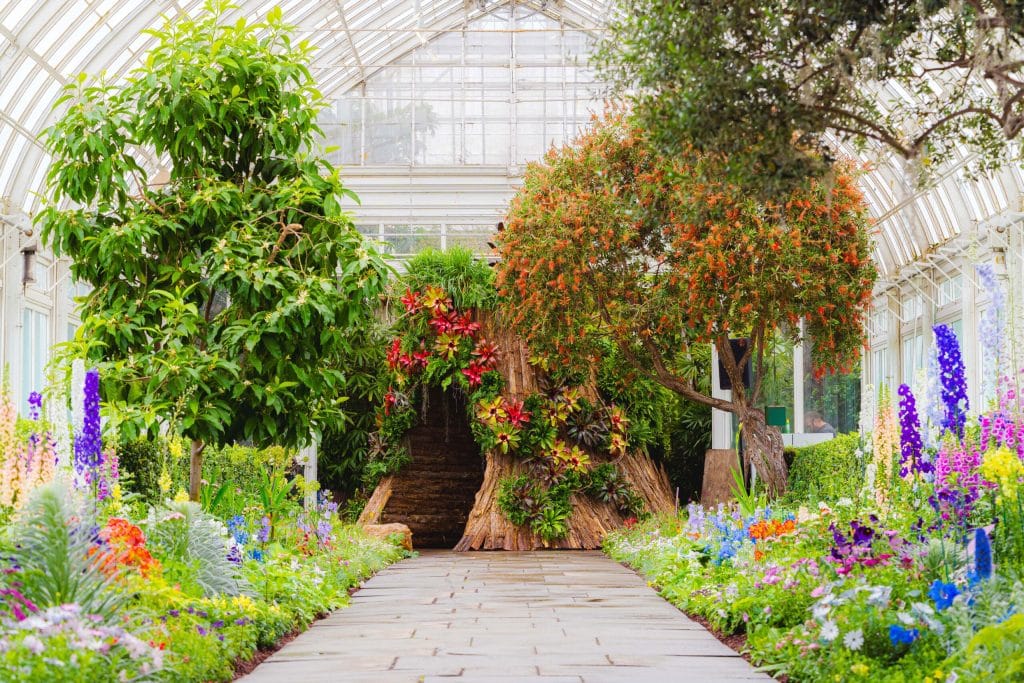 A photograph of a collection of brightly colored flowers and trees with a fake tree trunk in the middle under a glass ceiling as part of the "Wonderland: Curious Nature" exhibition at the New York Botanical Garden.