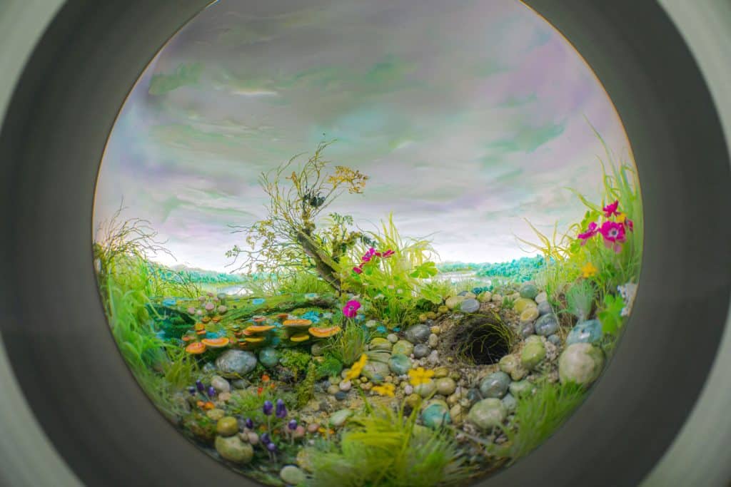 A photograph of a miniature landscape featuring flowers, pebbles, mushrooms, and a rabbit hole by artist Patrick Jacobs as part of the "Wonderland: Curious Nature" exhibition at the New York Botanical Garden.