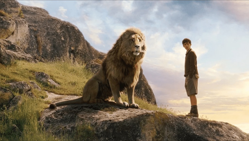 Still image from the 2005 high fantasy film "The Chronicles of Narnia: The Lion, the Witch, and the Wardrobe" featuring a CGI render of Aslan and Skandar Keynes standing on a rocky outcropping on the side of a hill. 