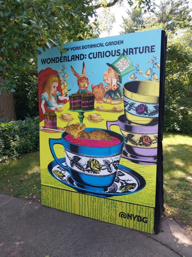 A photograph taken by Lynne Henderson of an advertisement for the New York Botanical Garden's "Wonderland: Curious Nature" exhibit featuring John Tenniel illustrations of Alice, the March Hare, and the Mad Hatter from "Alice's Adventures in Wonderland".