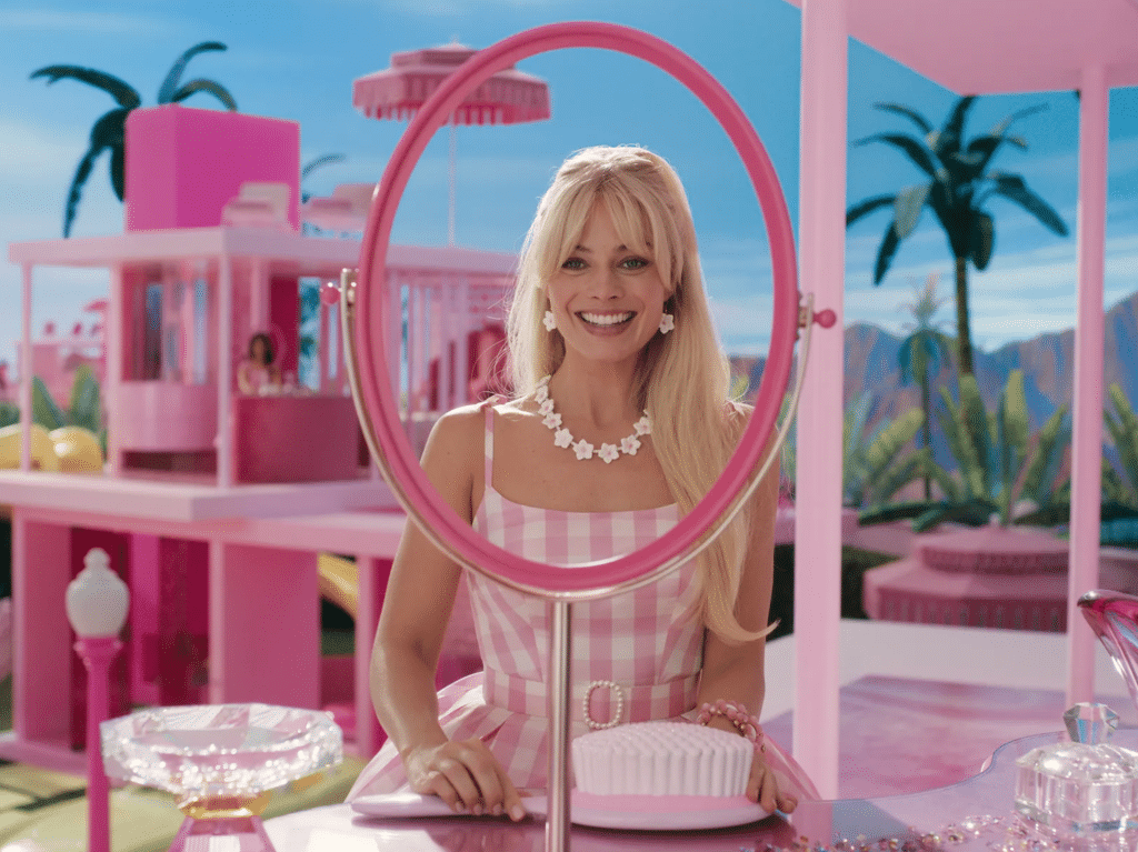 A screenshot from Greta Gerwig's 2023 film "Barbie," featuring Margot Robbie as the title character sitting at a desk with her face framed by a mirror frame. 