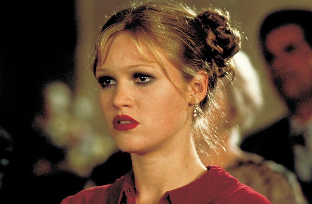 A still image from the 1998 mystery thriller film "Wicked" featuring Julia Stiles in a red top with dark eye shadow and her hair in a bun. 